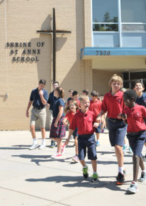 Children in red uniforms are walking and smiling outside Shrine of St. Anne School. A cross and school name are on the brick wall.